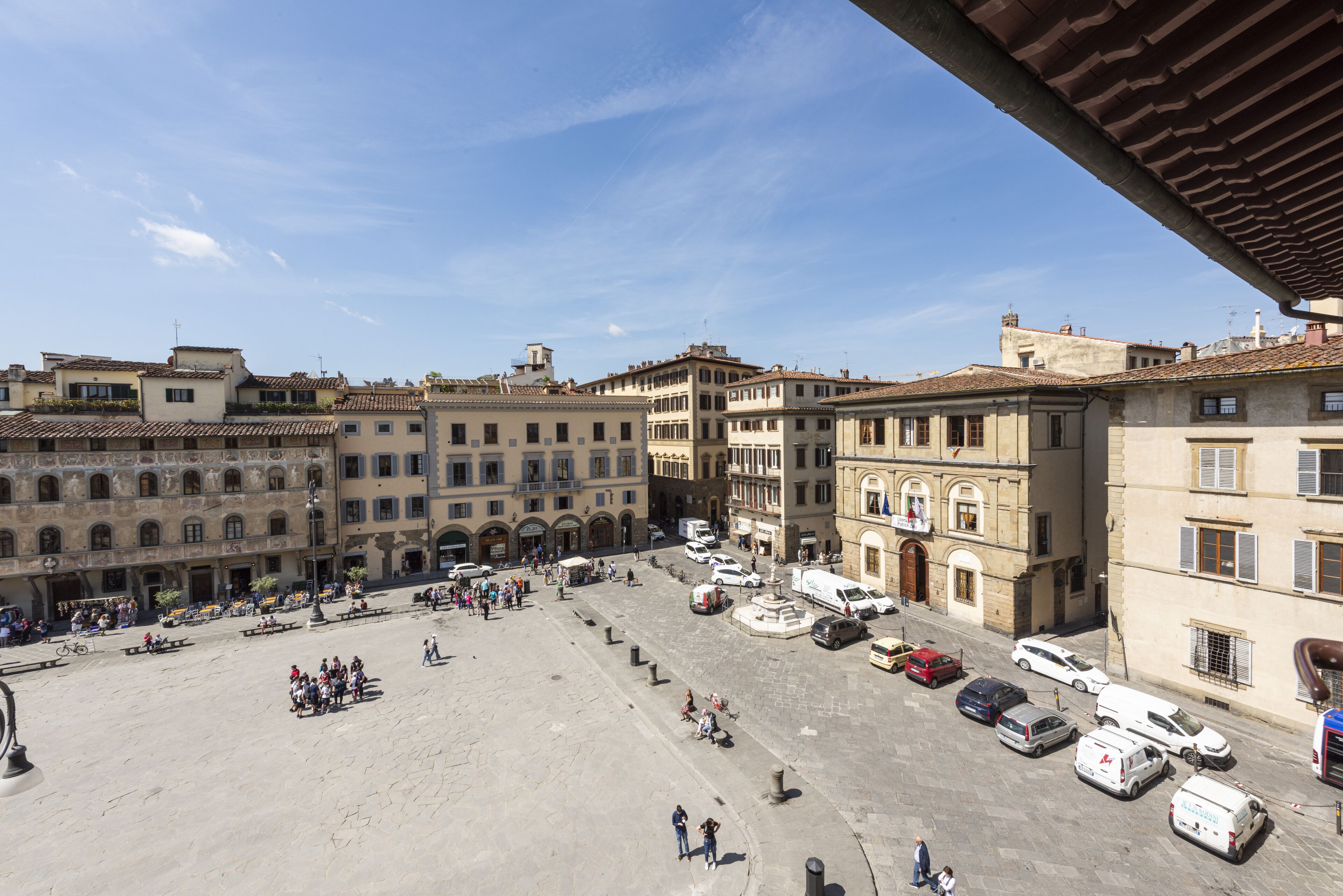 A spacious public square with historic buildings, a fountain, and a variety of parked vehicles.