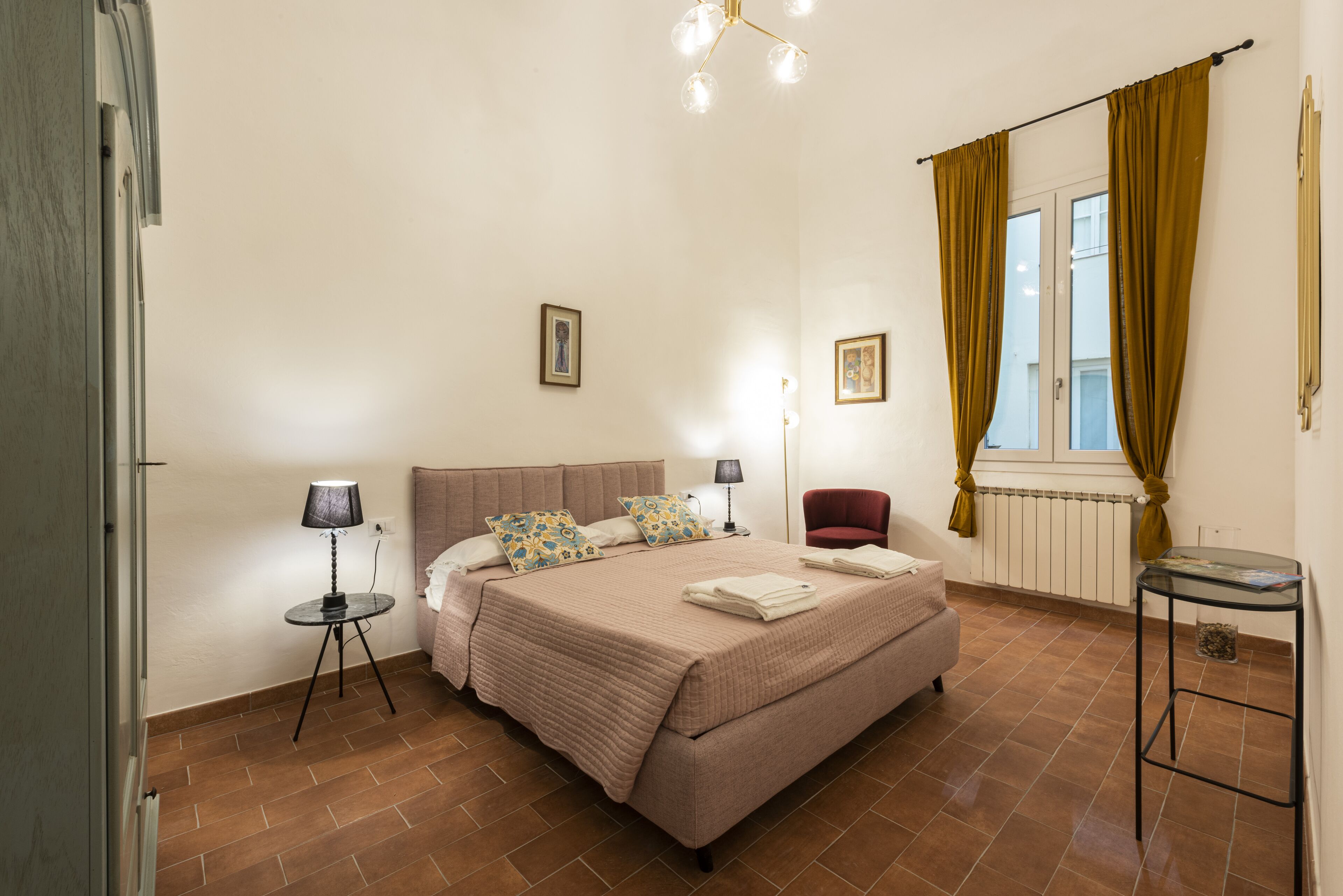 A bedroom with a bed, bedside lamps, a chair, a small table, and a window with curtains.