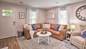 Un salon avec un canapé d’angle en cuir marron, une table basse blanche et un fauteuil beige. On y trouve une grande horloge murale, un tableau encadré et une petite table d’appoint avec une lampe.