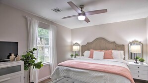 Une chambre à coucher comprenant un lit, des tables de chevet, un ventilateur de plafond, une télévision, une plante et une fenêtre avec des rideaux.