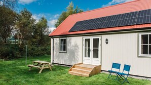 Een wit gebouw met een rood dak, zonnepanelen, een houten picknicktafel en blauwe klapstoelen.