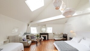 Una habitación acogedora y bien iluminada con techo inclinado, piso de madera y chimenea. Hay dos camas, una silla y una mesa con un jarrón de flores.