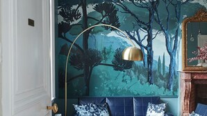 Una habitación con un gran mural de un bosque, un sofá azul, una lámpara de pie dorada y una chimenea clásica.