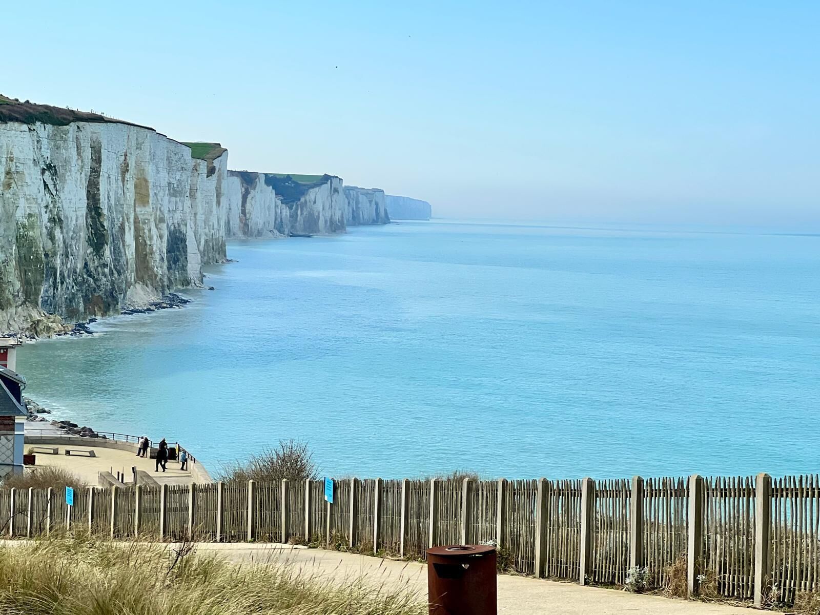 A coastal view with cliffs, a wooden fence, and a clear blue sea.