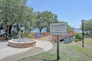 Un parque con un cartel dedicado a John J. Thomson, Jr., que presenta una estatua, una pasarela de madera y una fuente de piedra.