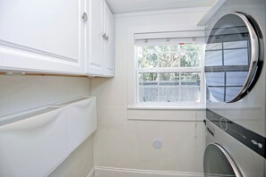 Un lavadero con lavadora de carga frontal, ventana con vista a árboles y muebles de cocina blancos.