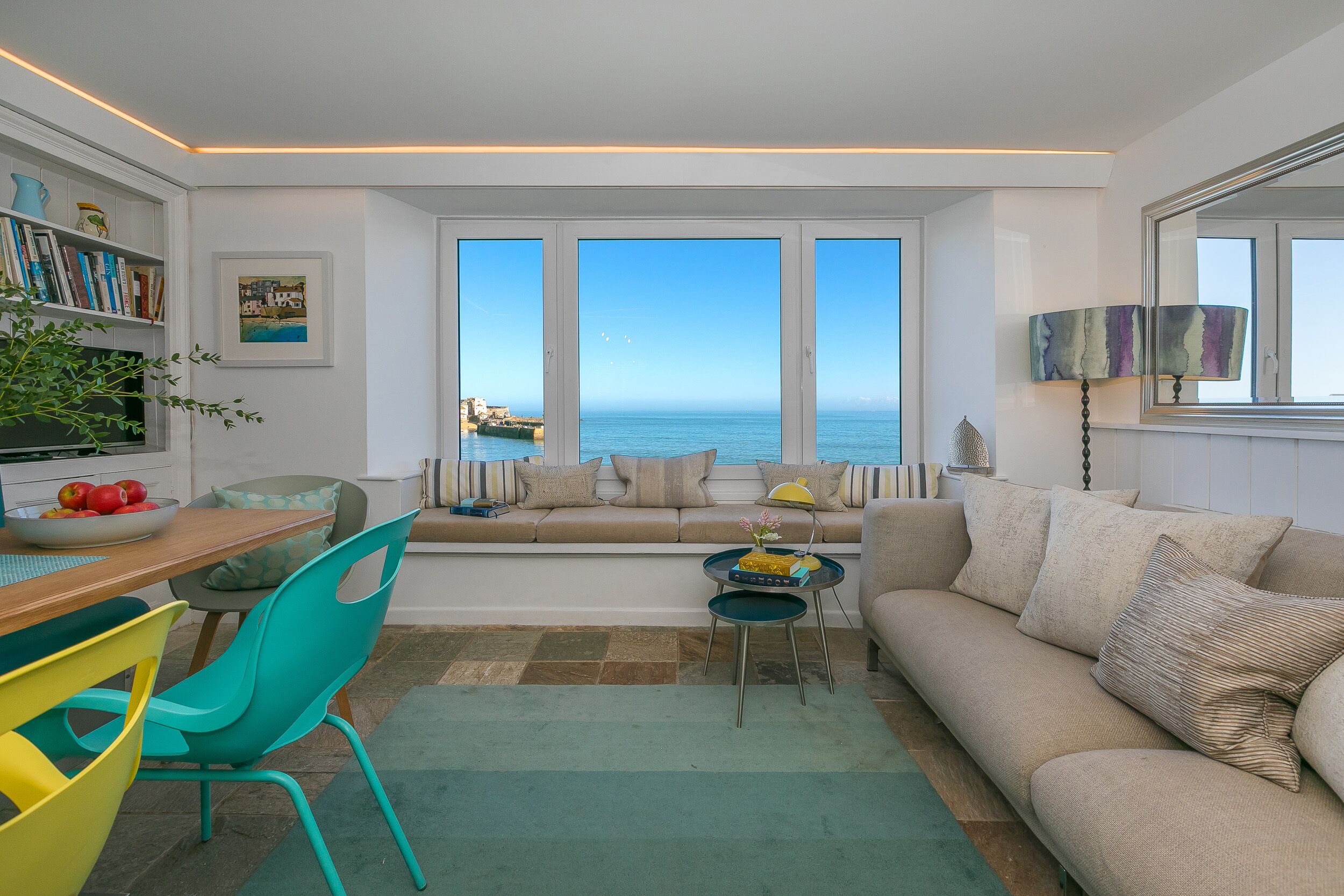 A modern living room with a large window overlooking the sea, a sofa, a dining table with chairs, and a bookshelf.
