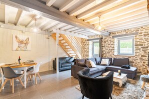 A spacious living area with a stone wall, a staircase, a dining table with chairs, and a black leather sofa. - Bel Even - Two Bedroom House, Sleeps 4 (Plouër-sur-Rance)