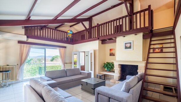 Ein geräumiger Wohnbereich mit einer großen Sofagarnitur, einem Couchtisch und einem Kamin. Eine Holztreppe führt auf eine obere Ebene, und es gibt einen Balkon mit Blick auf Grünflächen.