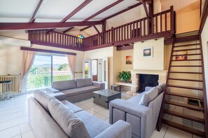 A spacious living area with a large sofa set, a coffee table, and a fireplace. A wooden staircase leads to an upper level, and there is a balcony with a view of greenery.