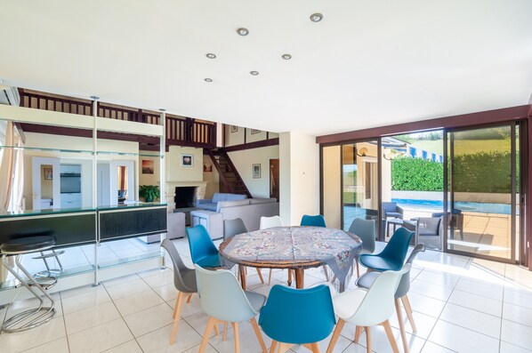 A spacious living area with a round table and chairs, a kitchen in the background, and a view of a swimming pool through large glass doors.