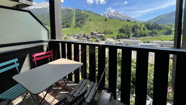 Ein Balkon mit Tisch und Stühlen, der auf eine gebirgige Landschaft mit Gebäuden und einer Eisenbahn blickt.