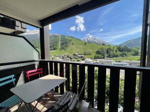 Un balcón con mesa y sillas, con vista a un paisaje montañoso que incluye edificios y una vía ferroviaria.