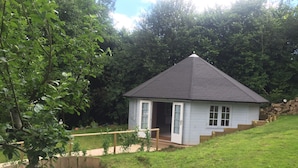 A small, single-story house with a dark roof and white exterior walls, featuring a front door and a small porch area. - Family Home With Large Garden and Pool Near Totnes (Totnes)