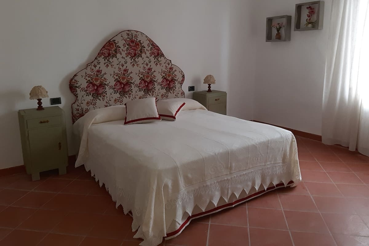 A bedroom with a floral headboard, a bed with white bedding and red trim, two bedside tables with lamps, and a tiled floor.
