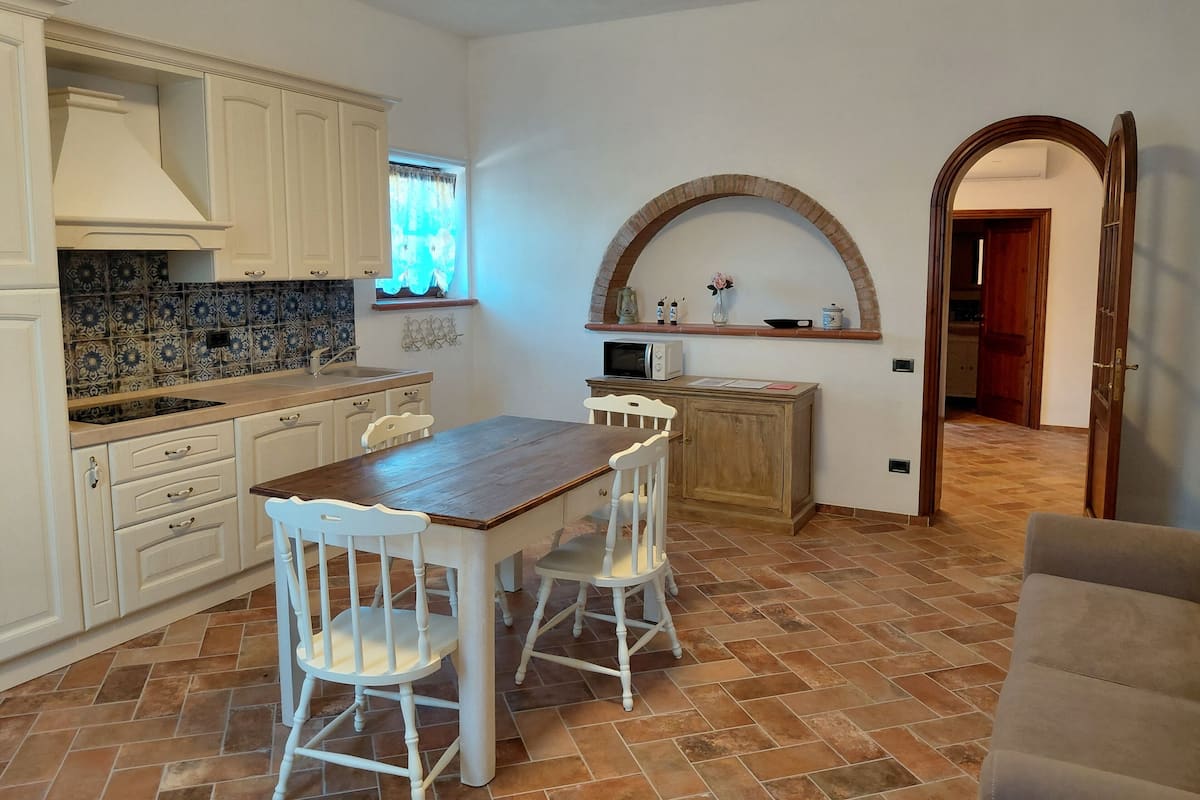 A kitchen with white cabinets, a wooden dining table with two chairs, a microwave, and a tiled backsplash.