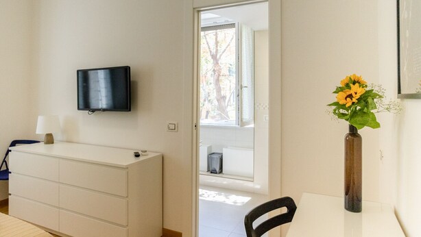 Habitación de hotel con televisor de pantalla plana en la pared, cómoda blanca, mesita con un jarrón de girasoles y una silla.