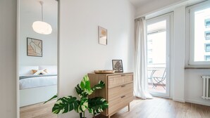 A modern room with a bed, a wooden dresser, a potted plant, and a large window with curtains. - Locarno City Apartments (Locarno)