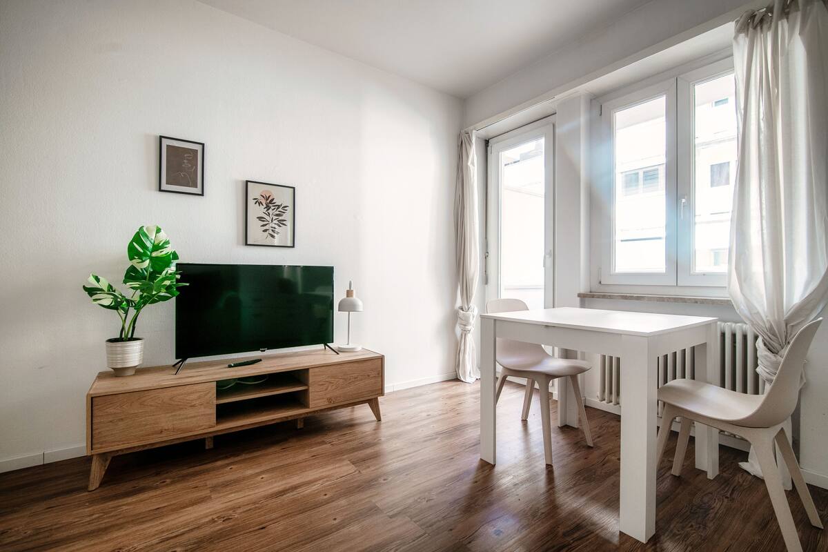 A modern living room with a wooden floor, a TV on a wooden stand, a dining table with chairs, and a large window with curtains.