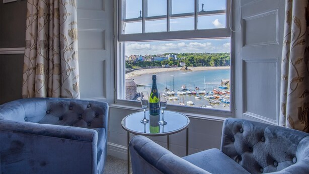 Una habitación con dos sillones capitonados, una mesita redonda con una botella y vasos, y un ventanal con vistas a una playa con barcos y edificios.