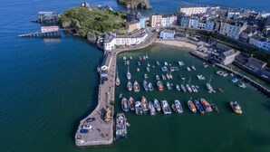 Vista aérea de un pueblo costero con un puerto deportivo lleno de barcos, un castillo en una colina y edificios coloridos a lo largo del paseo marítimo.