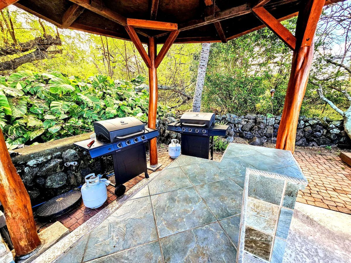 An outdoor barbecue area with two grills, a gas tank, and a stone-tiled floor.