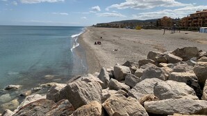 Une plage avec de gros rochers au premier plan, un ciel dégagé et des bâtiments en arrière-plan.