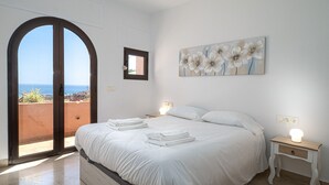 Une chambre à coucher avec un lit, une table de chevet, un tableau et une vue sur la mer, visible à travers une porte en arc de cercle.