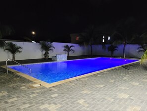 A swimming pool with a tiled deck, surrounded by palm trees and a white wall. - La Villetta, your home away from home (Willemstad)