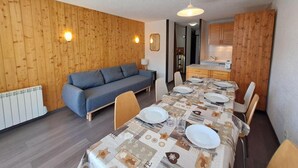 Et spiseområde med et langt bord dækket til fire personer, en sofa og et køkken i baggrunden.