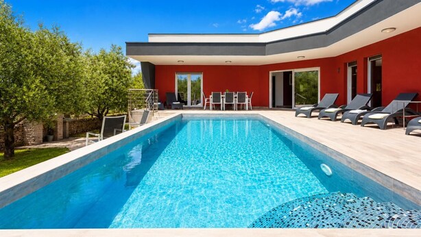 Een modern zwembadgedeelte met een helderblauw zwembad, ligbedden en een rood gebouw met een glazen deur.