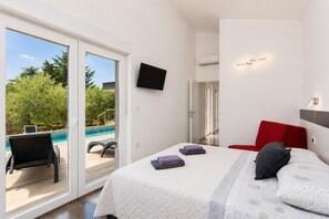 Un dormitorio con cama, televisor, una silla roja y vistas a una piscina al aire libre.