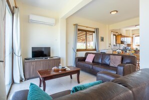 A living room with a sofa, a coffee table, and a TV. There is a kitchen in the background. - Blue Bay Villa Thea - Three Bedroom Villa, Sleeps 6 (Polis)