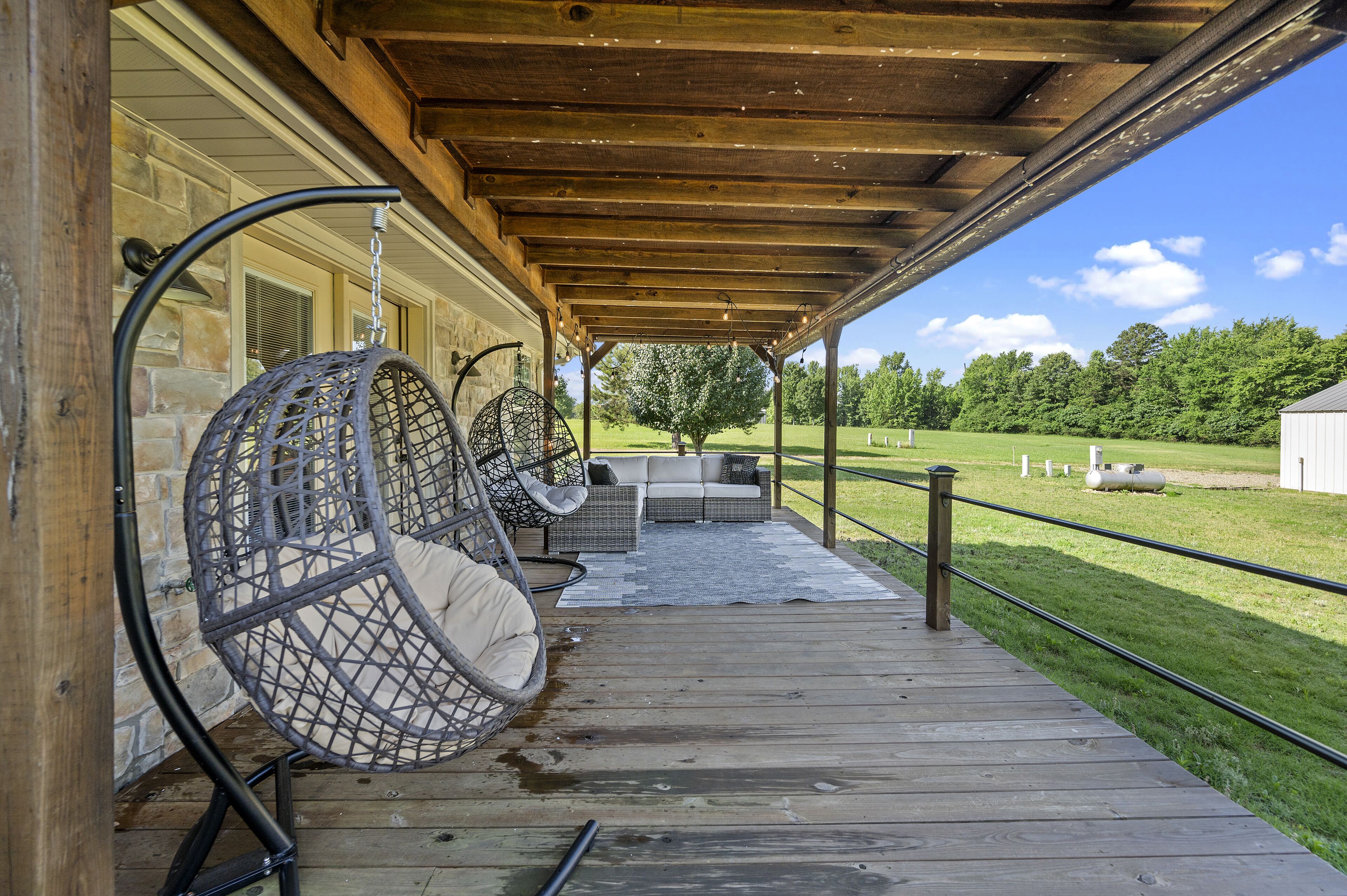 A covered outdoor seating area with wicker chairs and a sofa, a wooden deck, and a view of a green field.