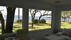 A room with large windows overlooking a grassy area and a body of water. There is a white sofa and a small table with a lamp. - Modern waterfront home on spacious wooded lot.  Sleeps 6.   (Elberta)