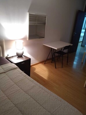 A bedroom with a bed, a bedside table with a lamp, a desk with a chair, and a window with blinds. - Quarto Partilhado (Queluz)