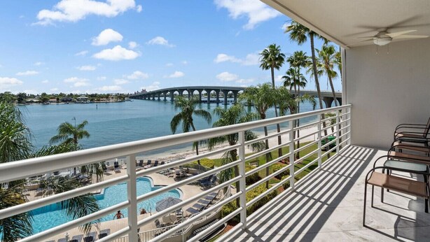 A balcony with a view of a beach, palm trees, a bridge, and a swimming pool.