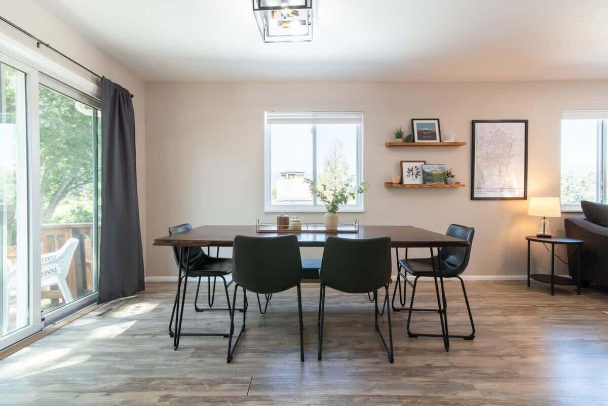 Un coin repas avec une table et des chaises, une fenêtre avec des rideaux, une plante posée sur le rebord de la fenêtre et une étagère murale agrémentée d'objets décoratifs.