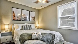 Une chambre à coucher bien rangée, avec un lit, deux tables de chevet, un ventilateur de plafond et une fenêtre avec des stores.