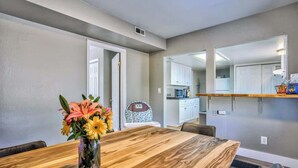 Un coin repas avec une table en bois, des chaises et un vase de fleurs. On aperçoit au fond une cuisine avec des meubles blancs et un four à micro-ondes.