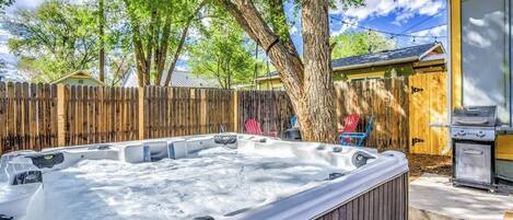 A hot tub in a backyard with a wooden fence, a barbecue grill, and a tree.