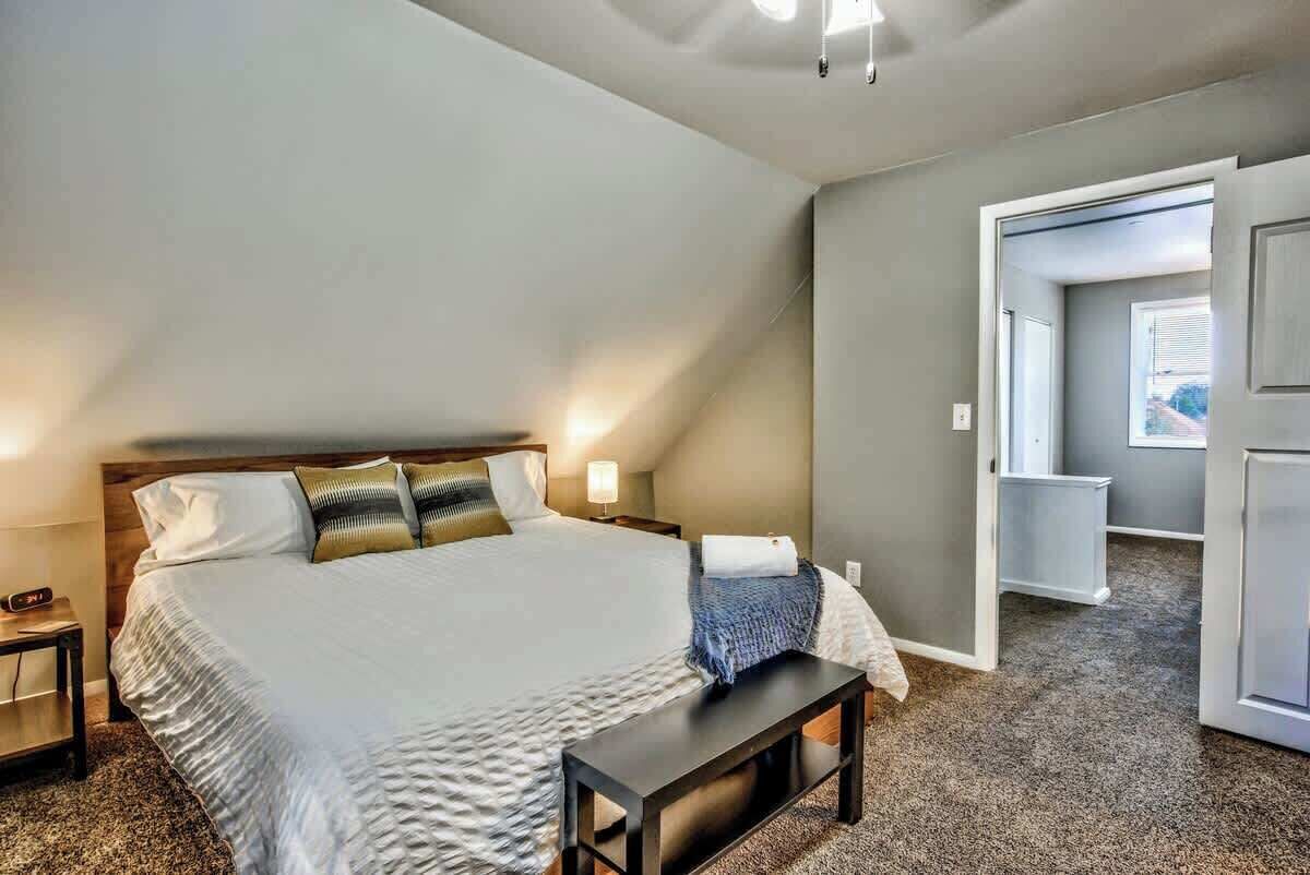 Quarto com uma cama grande, mesas de cabeceira, um banquinho e uma porta que dá para outro cômodo.