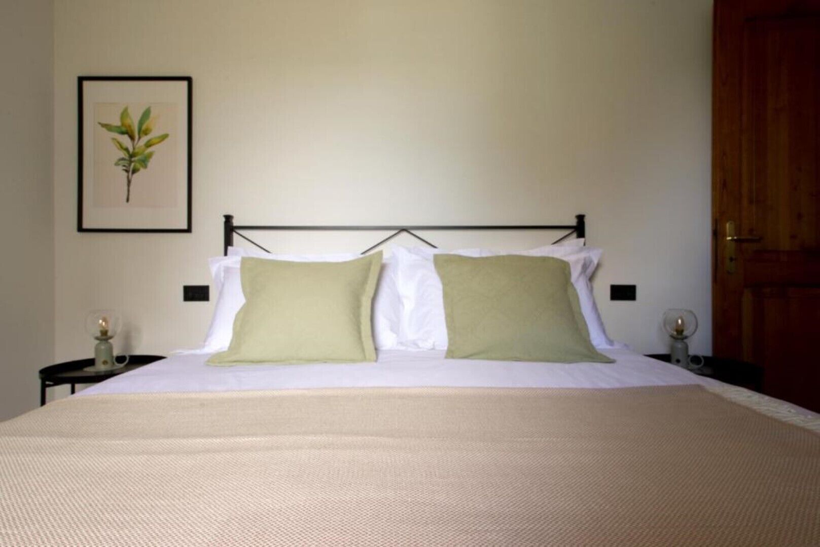A neatly made bed with two pillows, a framed plant on the wall, and a wooden door.