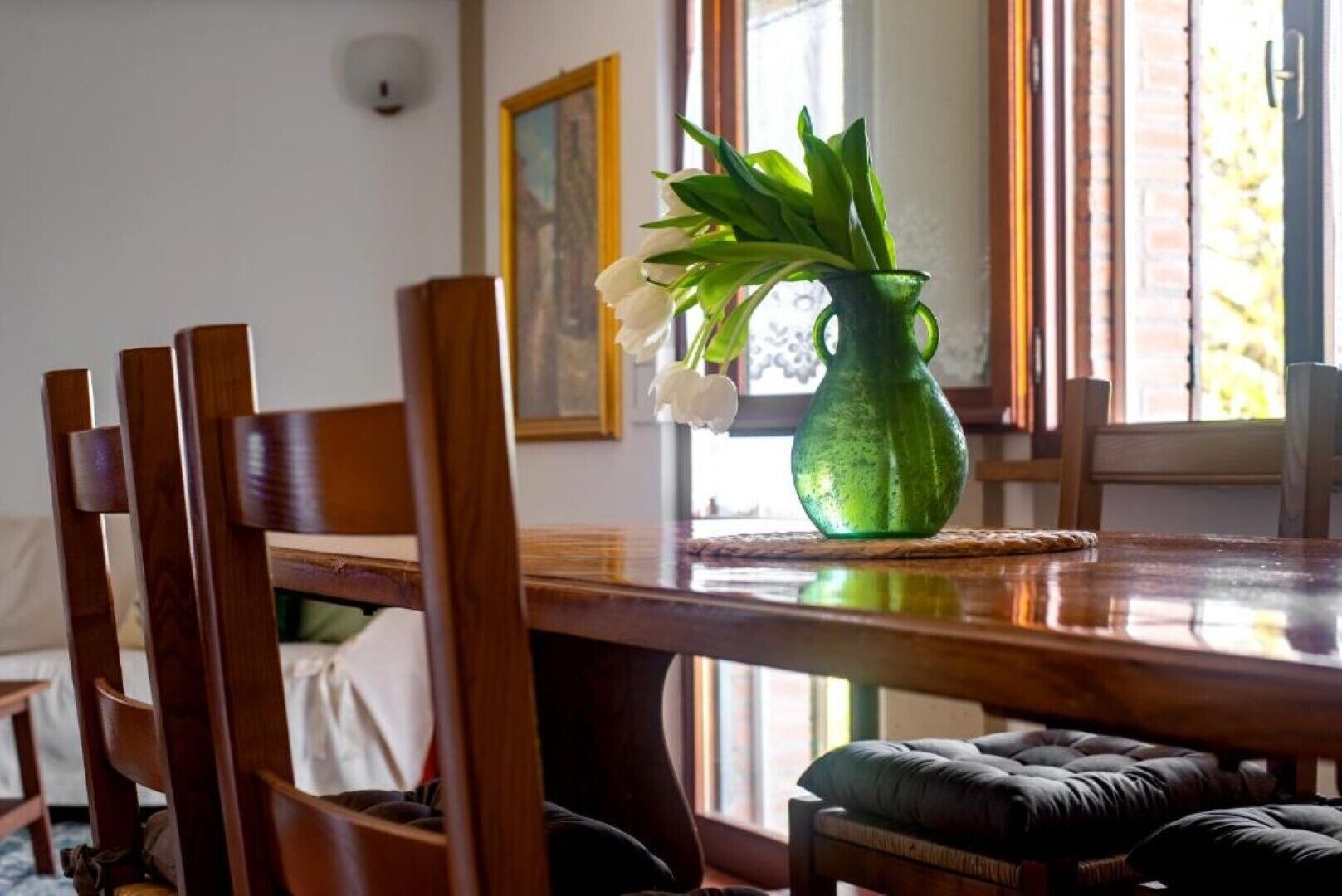 Une table à manger en bois sur laquelle est posé un vase vert contenant des fleurs blanches et des feuilles vertes.