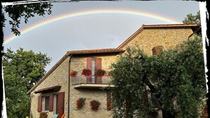 Edifici de pedra amb un balcó, envoltat de vegetació i un arc de set colors al cel.