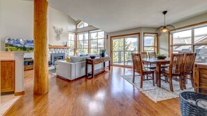 A spacious living area with a fireplace, a dining table with chairs, and large windows offering a view of the outdoors.