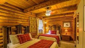 Une chambre de style rustique, aménagée dans une cabane en rondins, avec deux lits, des murs en bois, un ventilateur de plafond et une petite table de chevet avec une lampe.