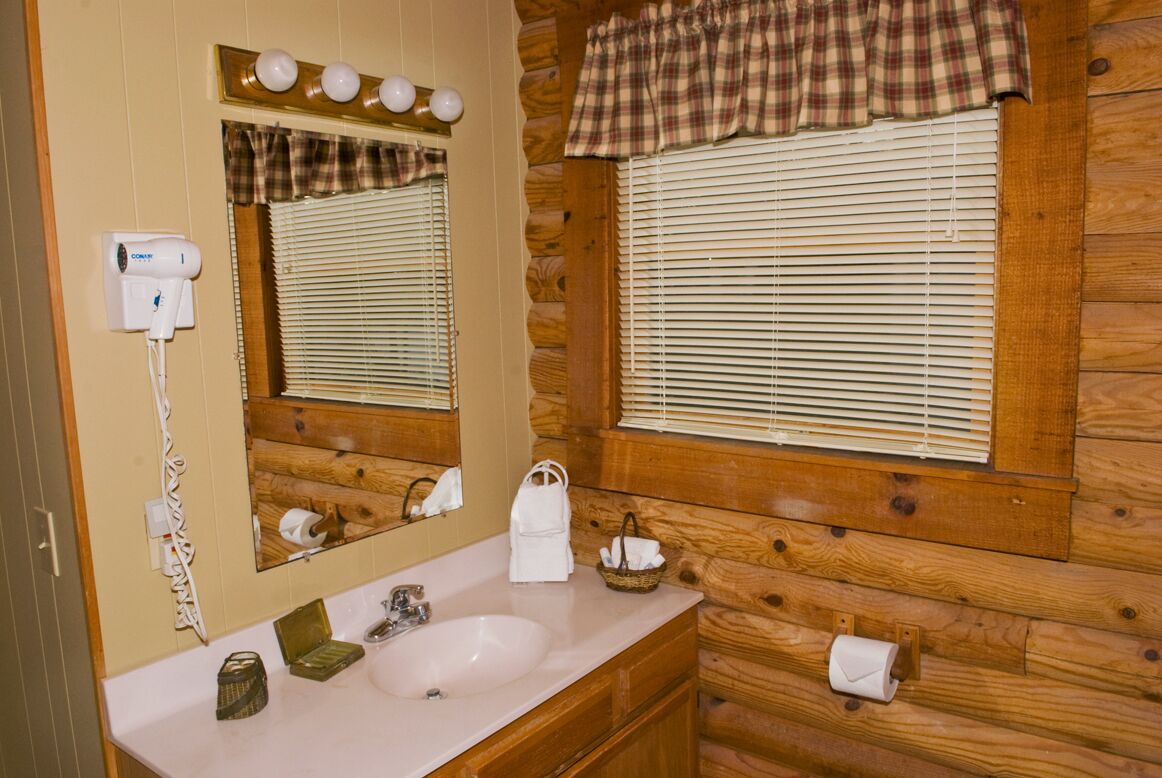 A wooden cabin bathroom with a sink, mirror, and window blinds.
