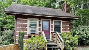 A small, single-story cabin with a red door, two windows with shutters, and a small porch with steps leading up to the door. - Queen Cozy Cabin on Main St (Blowing Rock)