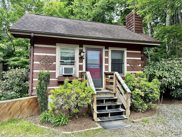 A wooden cabin with a red door, two windows, and a small porch with steps.